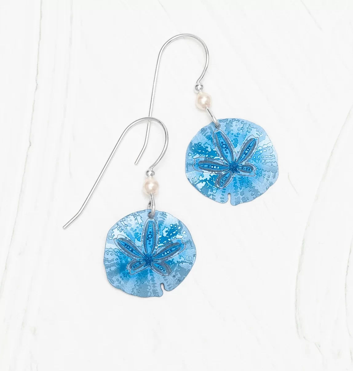 Explore ocean treasures with Holly Yashis Blue Sandra Shore Sand Dollar Earrings, showcasing intricate star details and pearl accents on hypo-allergenic silver hooks, displayed on a light textured background.