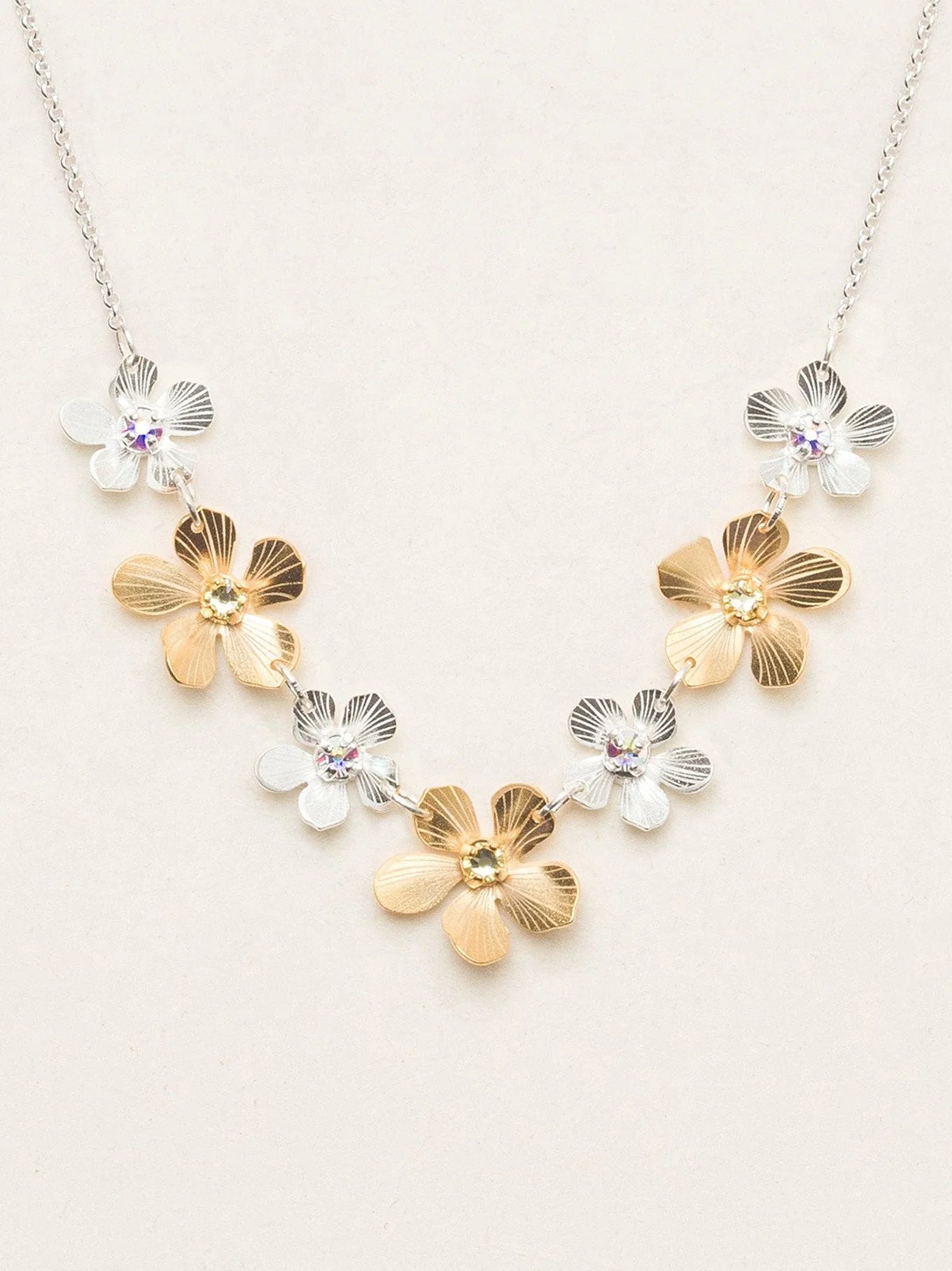 The Plumeria Classic Silver/Gold Necklace by Holly Yashi features alternating gold and silver flower-shaped charms with gemstone centers, beautifully displayed on a light beige background.