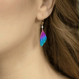 Close-up of a person wearing Holly Yashis Blue Violet Flutterby Monarch Earrings, featuring pink, purple, and blue feathers on an 18k gold-filled French wire. The delicate accessory contrasts with the persons flowing light brown hair.