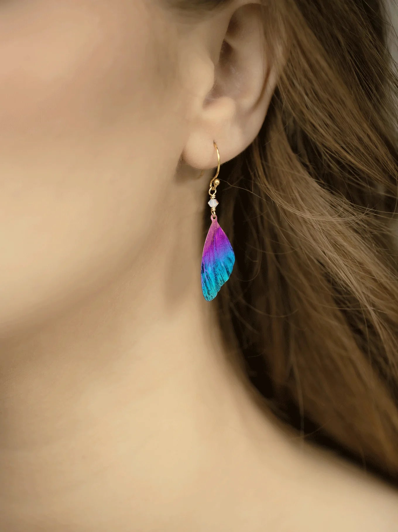 Close-up of a person wearing Holly Yashis Blue Violet Flutterby Monarch Earrings, featuring pink, purple, and blue feathers on an 18k gold-filled French wire. The delicate accessory contrasts with the persons flowing light brown hair.
