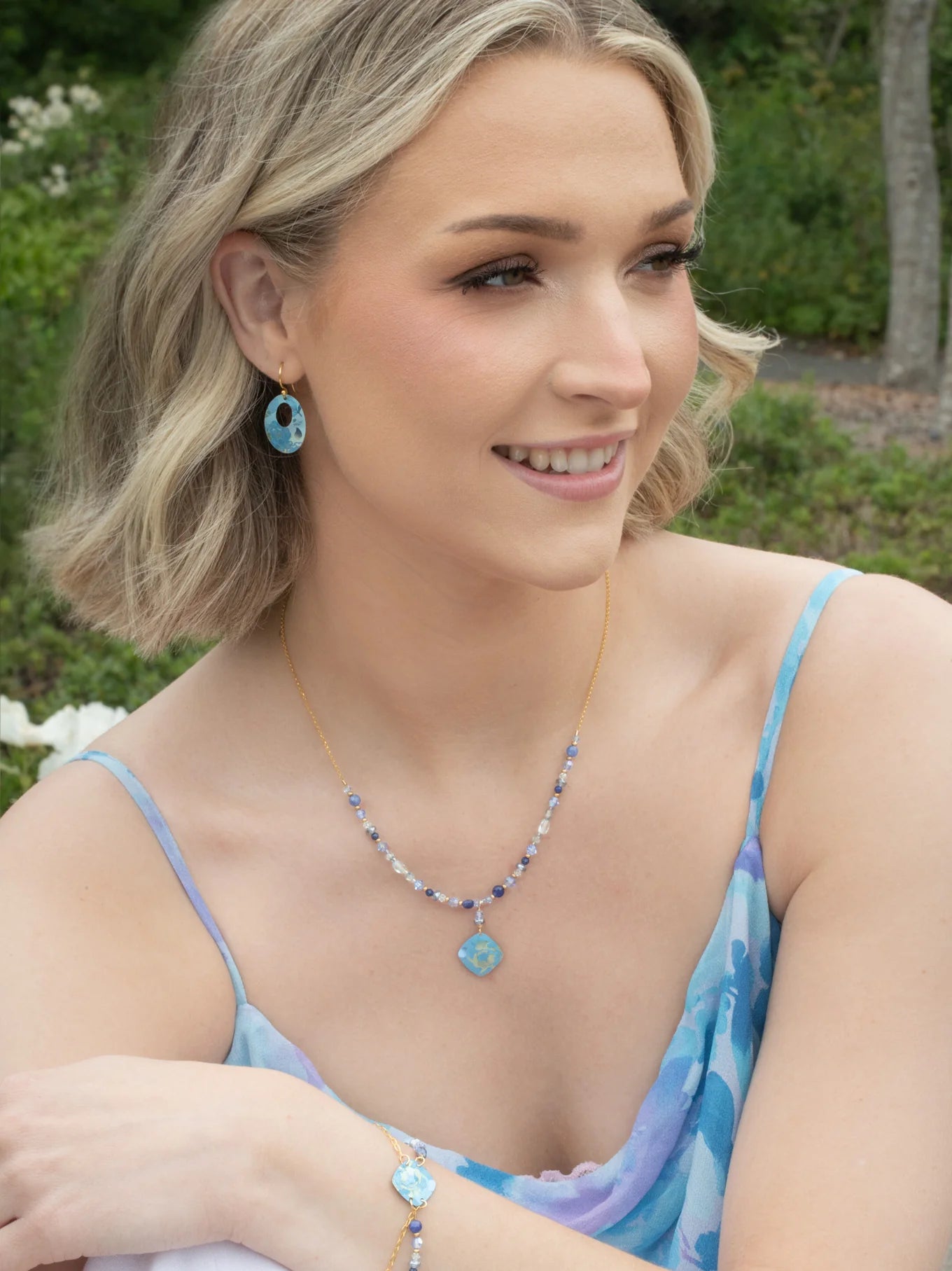 A woman with short blonde hair and natural makeup smiles outdoors, wearing the Holly Yashi Elena Beaded Necklace Violet Necklace, paired with matching earrings and bracelet; green foliage and flowers appear in the background.