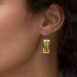 A close-up shows a person wearing Holly Yashis Teal Purple Eden Drop Earrings. These niobium earrings feature an intricate abstract leaf design in teal, blue, and yellow tones, dangling elegantly from gold hooks, beautifully contrasted against dark hair.
