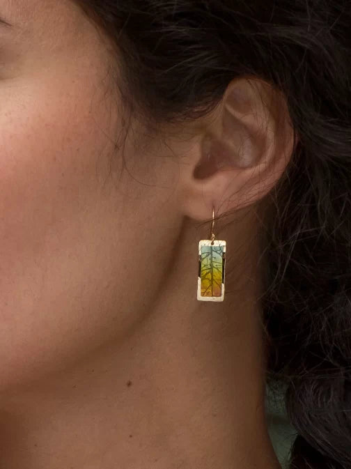 A close-up shows a person wearing Holly Yashis Teal Purple Eden Drop Earrings. These niobium earrings feature an intricate abstract leaf design in teal, blue, and yellow tones, dangling elegantly from gold hooks, beautifully contrasted against dark hair.
