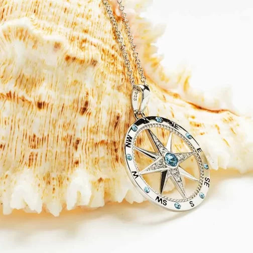 The Swarovski Crystals Aqua and White Compass Necklace by Shanore is beautifully presented on a large seashell. The sterling silver pendant, featuring a compass with dazzling blue gems, forms an exquisite accessory for any adventure.