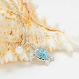 The Swarovski Crystals Aqua Turtle and Pearl Necklace by Shanore, featuring blue and clear crystals, elegantly drapes over a large seashell. A small pearl charm complements the turtle pendant against the softly focused textured seashell, enhancing the oceanic theme.