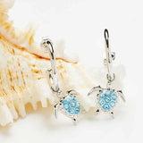 Shanores Ocean Jewelry collection features stunning Swarovski Crystals Post Hoop Dangle Turtle Earrings. These sterling silver hoops, adorned with small sparkling stones on the turtle shells, hang elegantly in front of a textured seashell backdrop.