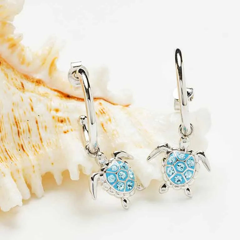 Shanores Ocean Jewelry collection features stunning Swarovski Crystals Post Hoop Dangle Turtle Earrings. These sterling silver hoops, adorned with small sparkling stones on the turtle shells, hang elegantly in front of a textured seashell backdrop.