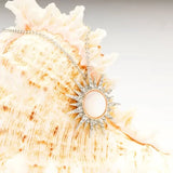 A Shanore Swarovski Crystals Mother of Pearl Rose Gold Sun Necklace is elegantly draped over a textured seashell, with its ornate sun pendant featuring a central oval stone surrounded by sparkling crystals against a softly blurred background.