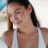 A woman with long dark hair is wearing a white tank top, blue butterfly-shaped earrings, and Shanores Swarovski Crystals Blue Sapphire Pearl Starfish Necklace. She is smiling slightly and looking down against a blurred beach backdrop.