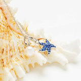 The Shanore Swarovski Crystals Blue Sapphire Pearl Starfish Necklace elegantly drapes over white coral, featuring a blue pendant adorned with shimmering crystals and a sapphire pearl charm against a light background.