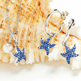 The Shanore jewelry set includes Swarovski Crystals Blue Sapphire Starfish Dangle Earrings and a matching necklace with a pearl pendant, all in sterling silver. These pieces shimmer beautifully against a textured seashell background.