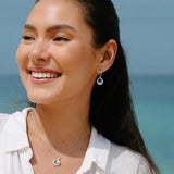 A woman with long dark hair smiles, wearing a white shirt, sterling silver earrings, and a pendant. Her Shanore Wave Earrings shimmer like Swarovski Crystals Sapphire/Aqua Post Hoop Ocean Jewelry before the blue ocean under a clear sky.