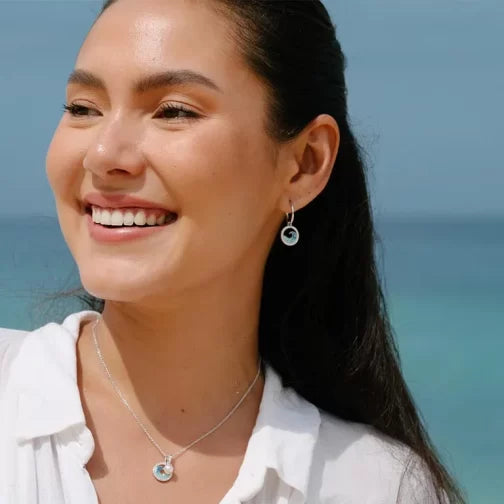 A smiling woman stands outdoors by the ocean, wearing a white shirt and Shanores Swarovski Crystals Blue Wave Drop Earrings. Her pendant necklace complements her look as her hair is pulled back against the serene sky and sea backdrop.