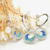 Silver jewelry displayed on a seashell backdrop, featuring a necklace with a pearl pendant. The Shanore Swarovski Crystals Sapphire/Aqua Post Hoop Wave Earrings showcase circular designs with sparkling blue and yellow patterns that shimmer like crystals.