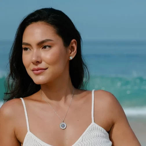 A woman with long dark hair stands by the beach, wearing a white tank top and a Shanore Swarovski Crystals Mother of Pearl Blue Sapphire Compass Necklace. The serene scene is completed by the ocean waves and blue sky behind her.