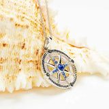 The Shanore Swarovski Crystals Mother of Pearl Blue Sapphire Compass Necklace features a pendant with cardinal direction letters and a central blue gem, glimmering alongside a textured seashell, all elegantly set on a thin silver chain.