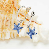 Shanores Swarovski Crystals Blue Sapphire Starfish Dangle Earrings are elegantly displayed against a white, textured shell background. The starfish charms sparkle with small blue gems, adding a touch of ocean-inspired elegance to each piece.