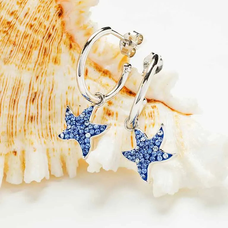 Shanores Swarovski Crystals Blue Sapphire Starfish Dangle Earrings are elegantly displayed against a white, textured shell background. The starfish charms sparkle with small blue gems, adding a touch of ocean-inspired elegance to each piece.