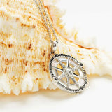 The Shanore Swarovski Crystals White Compass Necklace, adorned with a sterling silver compass rose and central crystal, is elegantly displayed on a textured seashell backdrop.