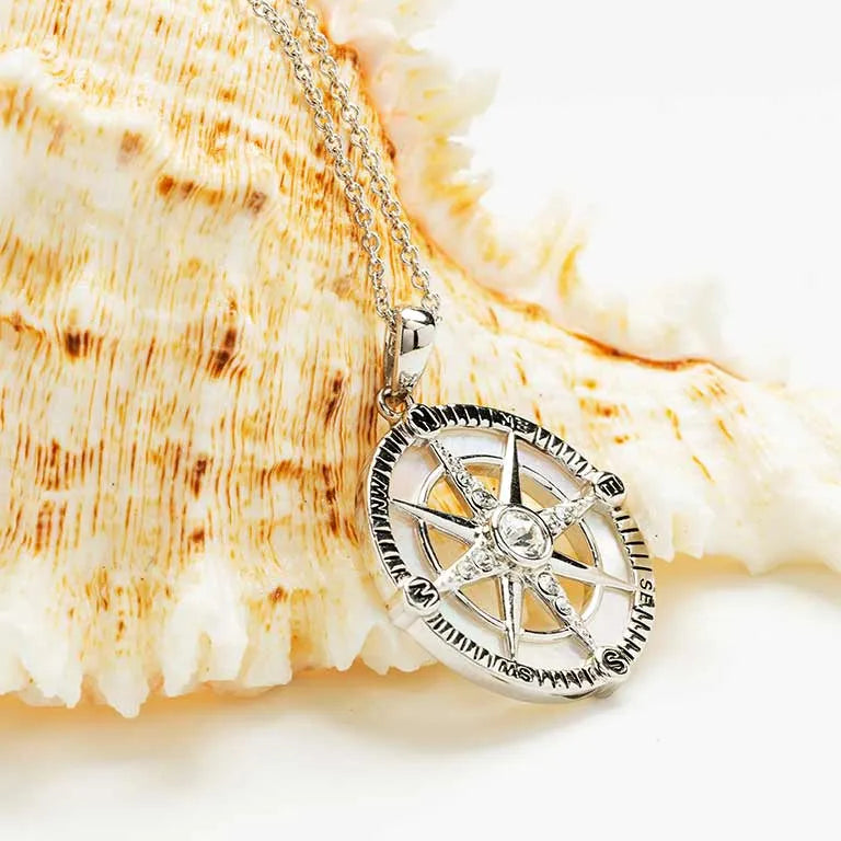 The Shanore Swarovski Crystals White Compass Necklace, adorned with a sterling silver compass rose and central crystal, is elegantly displayed on a textured seashell backdrop.