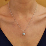 A person wears the Shanore Swarovski Crystals Medium Aqua Starfish Necklace-Ocean Jewelry, adorned with aquamarine crystals, against a blurred background. The jewelry stands out against the persons skin, with a dark top visible at the shoulders.