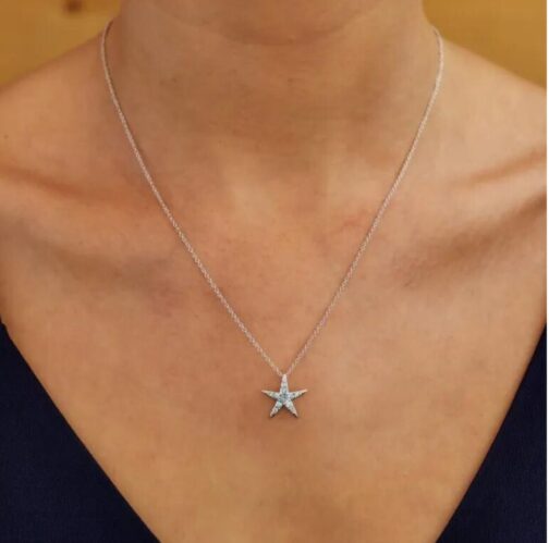 A person wears the Shanore Swarovski Crystals Medium Aqua Starfish Necklace-Ocean Jewelry, adorned with aquamarine crystals, against a blurred background. The jewelry stands out against the persons skin, with a dark top visible at the shoulders.