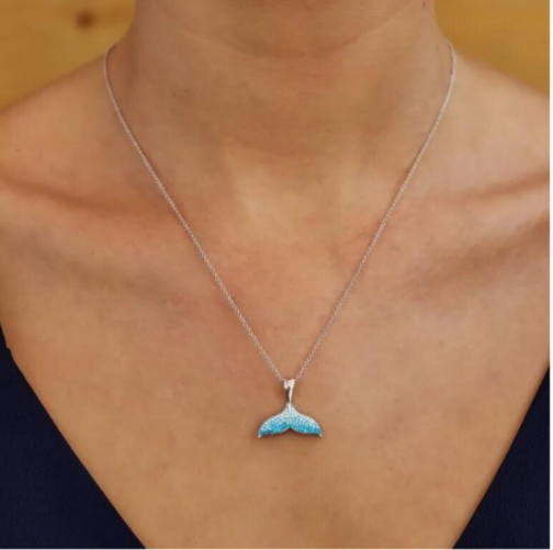 A person wears a Swarovski Crystals Aqua Mermaid Tail Necklace by Shanore, featuring a turquoise mermaid tail pendant on a sterling silver chain, set against a neutral backdrop. They wear a dark V-neck top.