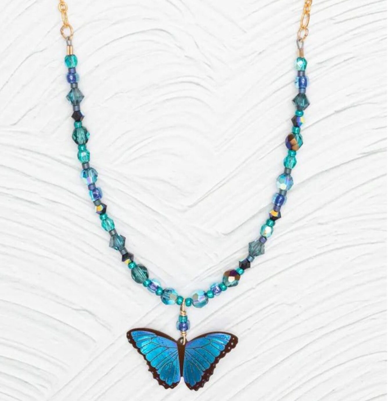 Discover the Blue Bella Butterfly Beaded Necklace by Holly Yashi, a handcrafted piece with a vibrant blue butterfly pendant surrounded by blue and teal beads, all beautifully set against a textured white backdrop.