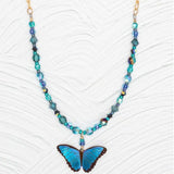 Discover the Blue Bella Butterfly Beaded Necklace by Holly Yashi, a handcrafted piece with a vibrant blue butterfly pendant surrounded by blue and teal beads, all beautifully set against a textured white backdrop.