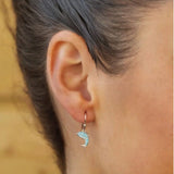 Close-up of a persons ear wearing Shanores Swarovski Crystals Aqua Cry Dolphin Drop Earrings, Ocean Jewelry. The earrings feature dolphin shapes with turquoise-like accents, while the background is softly blurred in warm tones.