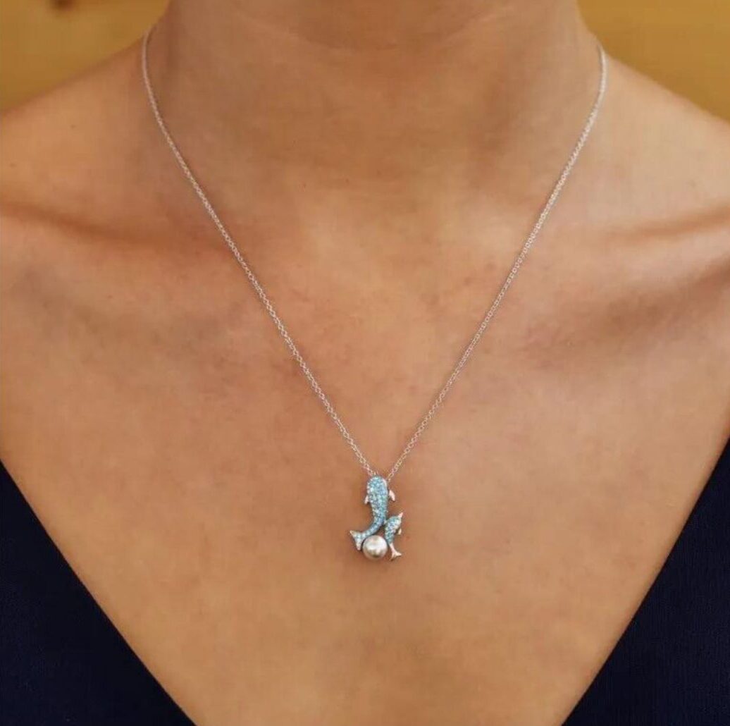A woman dazzles in a Shanore Swarovski Crystals Dolphin and Baby Pearl Necklace, her V-neck top enhancing the ocean-inspired elegance against a warm, neutral backdrop.