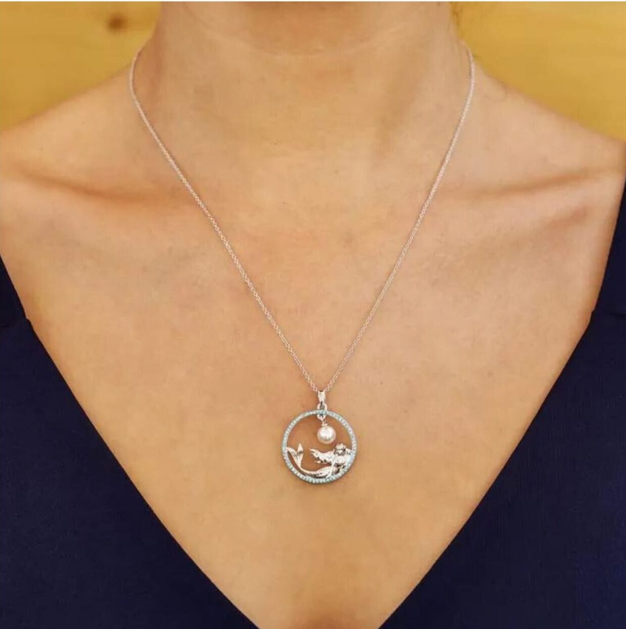 Close-up of a Shanore Swarovski Crystals Circle Mermaid with Pearl Necklace-Ocean Jewelry, featuring a mermaid pendant encircled delicately. The thin chain sparkles with Swarovski Crystals against a warm-toned backdrop, worn over a dark V-neck top.