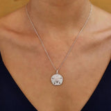 A person is wearing a Shanore Swarovski Crystals White Sand Dollar Necklace, featuring a skull design. Dressed in a black V-neck top, the focus is on how the Swarovski crystals accentuate the jewelry against their skin and fabric.