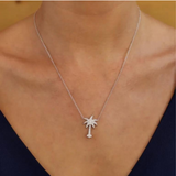 A person elegantly wears Shanore’s Swarovski Crystals White Palm Tree Necklace-Ocean Jewelry with a diamond-adorned sterling silver pendant, styled over a dark blue V-neck. A warm, neutral backdrop amplifies the timeless sophistication.