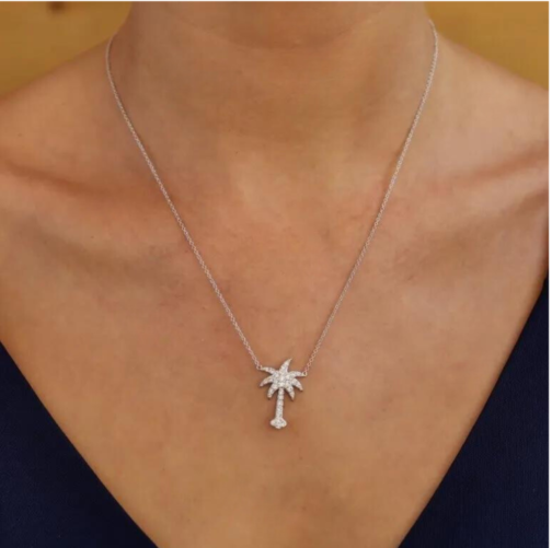 A person elegantly wears Shanore’s Swarovski Crystals White Palm Tree Necklace-Ocean Jewelry with a diamond-adorned sterling silver pendant, styled over a dark blue V-neck. A warm, neutral backdrop amplifies the timeless sophistication.