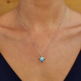 Someone wearing the elegant Shanore Swarovski Crystals Medium Turtle Aqua Necklace-Ocean Jewelry with a shimmering pendant, featuring a textured light blue center adorned with delicate details. The background is softly blurred, and the person wears a dark V-neck top.