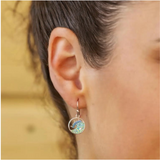 Close-up of a persons ear adorned with Swarovski Crystals Blue Wave Earrings-Ocean Jewelry by Shanore, showcasing turquoise gemstones in a wave pattern on sterling silver. A blurred wooden background adds warmth to the elegant design.
