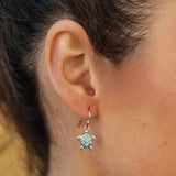 A close-up reveals a turtle-shaped earring, crafted in sterling silver with a blue textured center and Swarovski crystals. The earring, from Shanores Swarovski Crystals Aqua Drop Turtle Earrings - Ocean Jewelry collection, shines beautifully against the persons lightly tanned skin and blurred background.