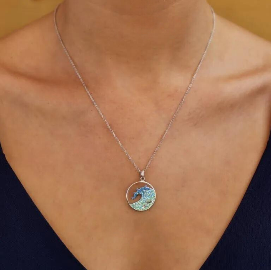 A close-up reveals a person wearing the Swarovski Crystals Blue Wave Necklace by Shanore. This sterling silver piece showcases a blue dolphin leaping over crystalline waves on a shimmering blue and green backdrop, complementing their dark V-neck top beautifully.