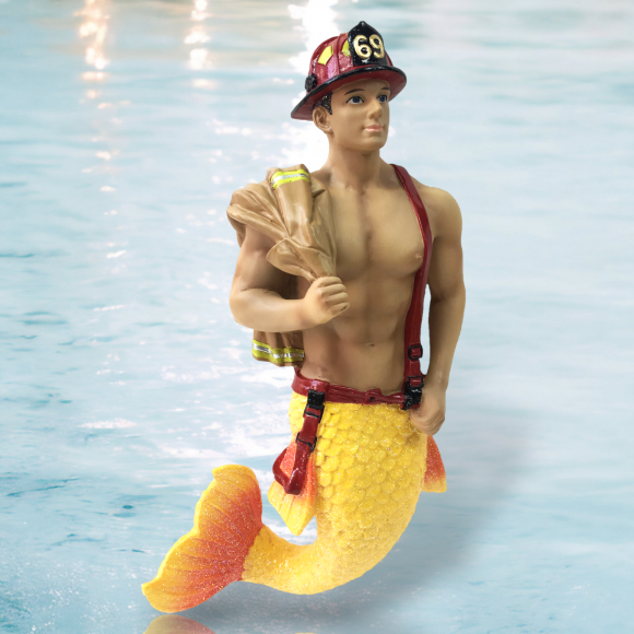 The Back Draft Ornament by December Diamonds features a merman with a muscular upper body and vibrant yellow fish tail, wearing a firefighters helmet and suspenders, confidently holding a hose against a water backdrop.