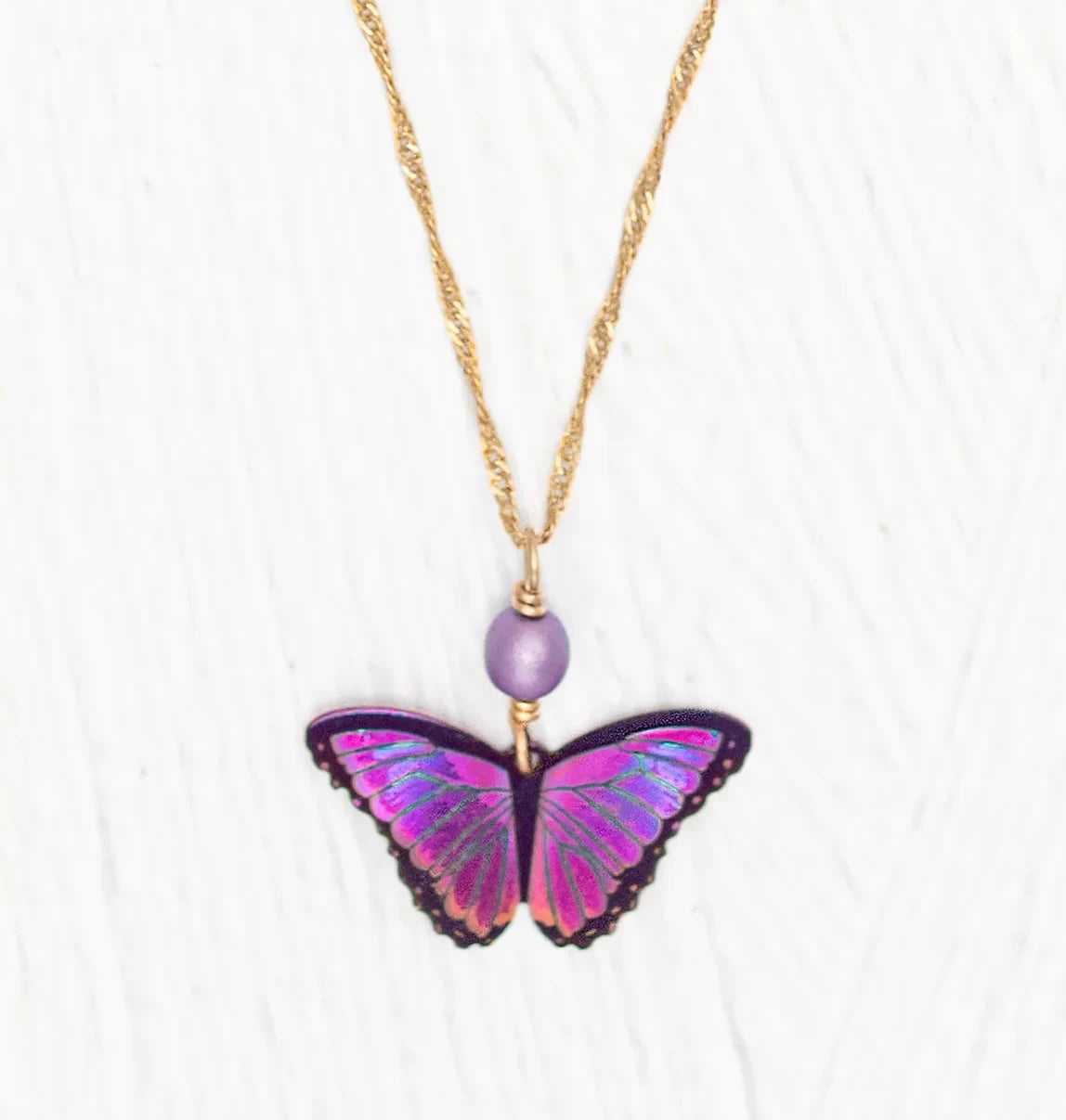 Presenting the Ultra Violet Bella Butterfly Necklace by Holly Yashi, featuring a detailed purple and pink butterfly pendant and a niobium bead, all elegantly hanging on an 18kt gold-plated twisted chain against a white textured background.