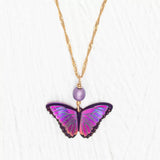 Presenting the Ultra Violet Bella Butterfly Necklace by Holly Yashi, featuring a detailed purple and pink butterfly pendant and a niobium bead, all elegantly hanging on an 18kt gold-plated twisted chain against a white textured background.