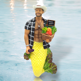 The Big N Tasty Ornament by December Diamonds features a mermaid with a yellow tail and cowboy hat, standing in shallow water. Wearing a plaid shirt, she carries a Farm Fresh basket with an eggplant—ideal for adding whimsical flair to your holiday decor.