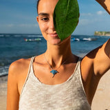 A woman on a beach, smiling, holds a large green leaf by her face. She wears a beige knitted tank top and displays a Shanore Swarovski Crystals Blue Stingray Necklace. The sea and distant shoreline appear under a clear blue sky in the background.