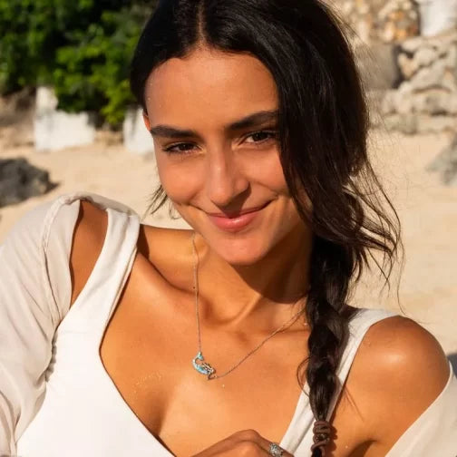 A woman with dark braided hair wearing a Shanore Swarovski Crystals Aqua Mermaid Necklace smiles at the camera. Shes in a white top enjoying a sunny beach day, surrounded by greenery and rocks.