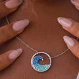 Manicured nails touch a Shanore Swarovski Crystals Blue Wave Necklace from Ocean Jewelry, its sterling silver circular pendant gleaming. A matching wave-patterned ring adorned with Swarovski Crystals graces the finger against a beautifully blurred background.