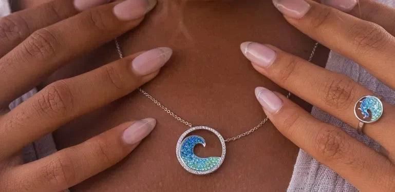 Manicured nails touch a Shanore Swarovski Crystals Blue Wave Necklace from Ocean Jewelry, its sterling silver circular pendant gleaming. A matching wave-patterned ring adorned with Swarovski Crystals graces the finger against a beautifully blurred background.