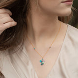 A person wears a light beige blouse and the Blue Picaflor Necklace by Holly Yashi, featuring a small colorful bird pendant on an adjustable sterling silver chain. Their hand grazes the necklace as long brown hair flows softly in front of a gently blurred background.