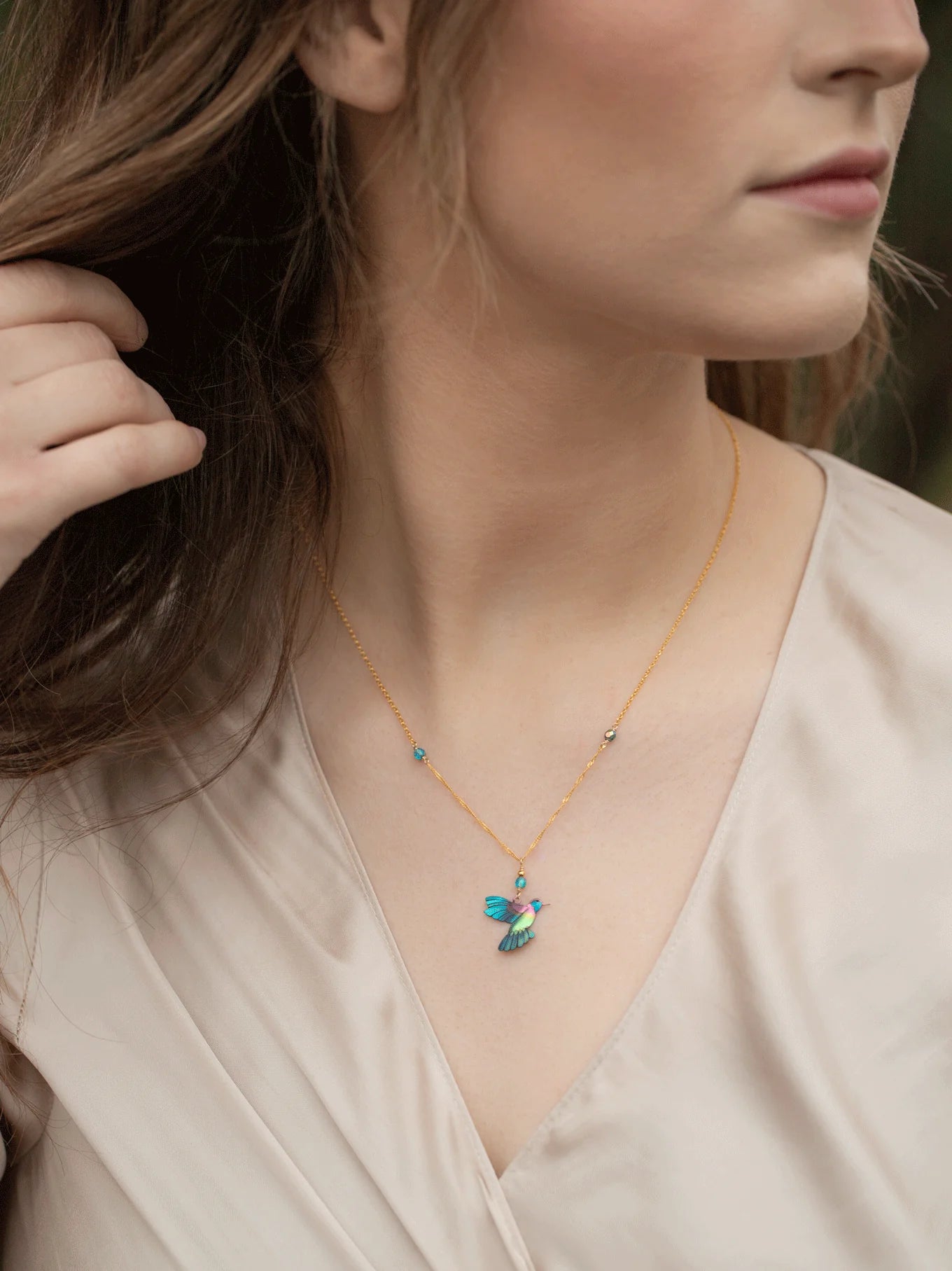 A person wears a light beige blouse and the Blue Picaflor Necklace by Holly Yashi, featuring a small colorful bird pendant on an adjustable sterling silver chain. Their hand grazes the necklace as long brown hair flows softly in front of a gently blurred background.