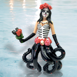 The Catrina Ornament by December Diamonds features a skeletal face with a corset adorned in roses and a small skull, complemented by black octopus tentacles. It holds red roses and wears a rose headdress, all hand-painted against a light blue background.